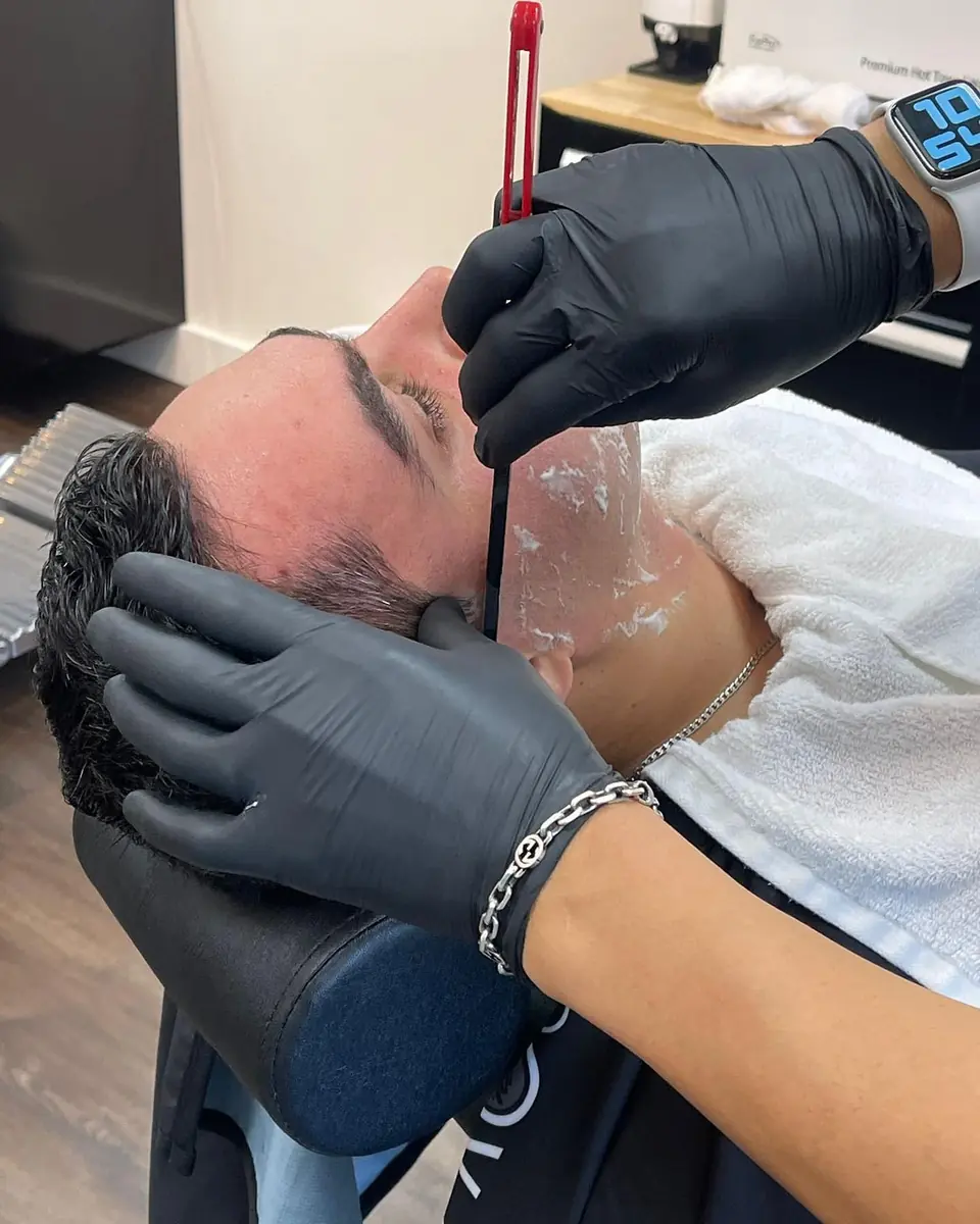 Gloved hands shaving a lathered cheek with a straight razor